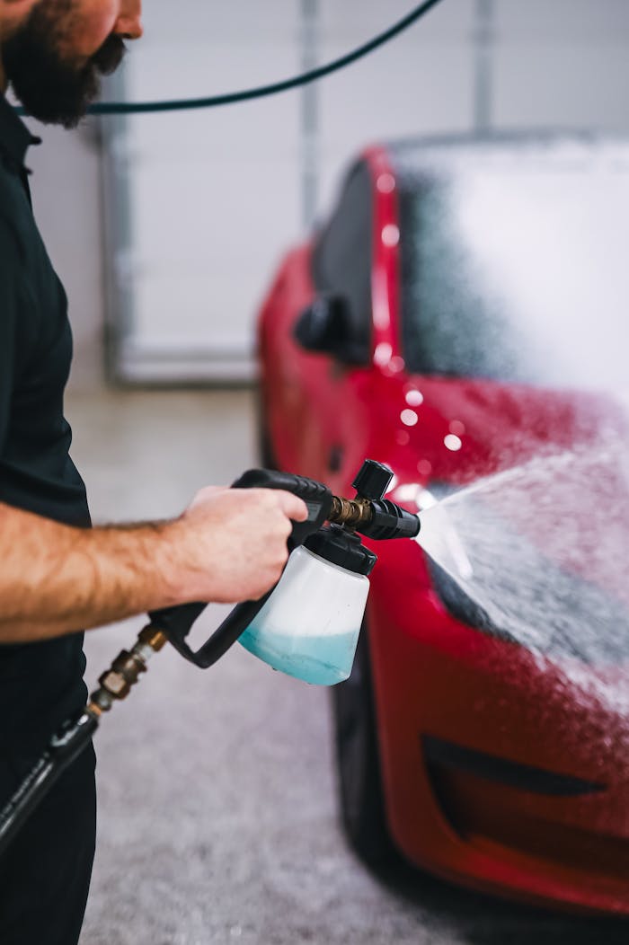 A man uses a foam sprayer to clean a red car indoors, showcasing car detailing.